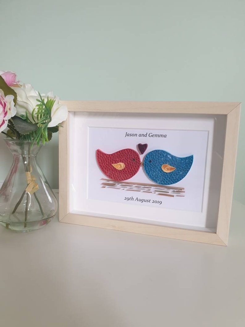 May include: A framed paper craft of two birds kissing with a heart above them. The birds are red and blue with yellow accents. The frame is light wood and the text reads "Jason and Gemma 29th August 2019".