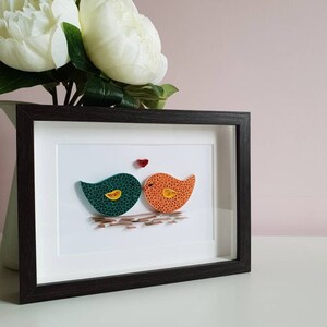 May include: A framed paper craft artwork featuring two birds in green and orange colors, kissing with a small red heart above them.
