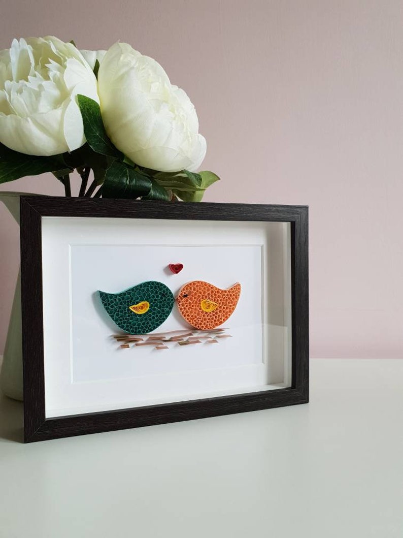 May include: A framed paper craft artwork featuring two birds in green and orange colors, kissing with a small red heart above them.