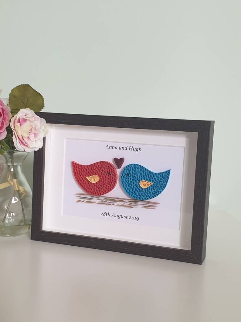 May include: A framed paper craft artwork featuring two birds, one red and one blue, with a small heart above them. The text "Anna and Hugh" and "28th August 2019" is printed below the birds.