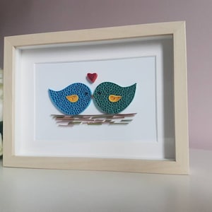 May include: A framed paper craft artwork featuring two blue and green birds kissing with a red heart above them. The birds are perched on a brown and gold nest.
