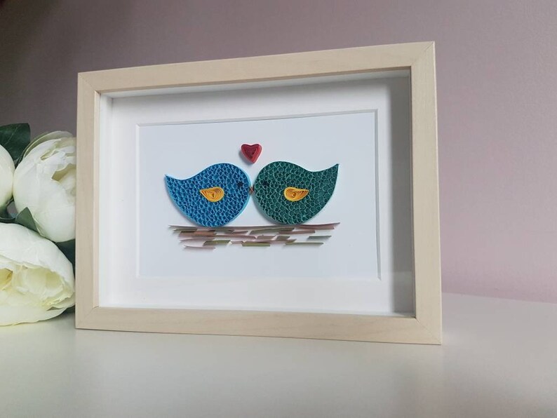 May include: A framed paper craft artwork featuring two blue and green birds kissing with a red heart above them. The birds are perched on a brown and gold nest.