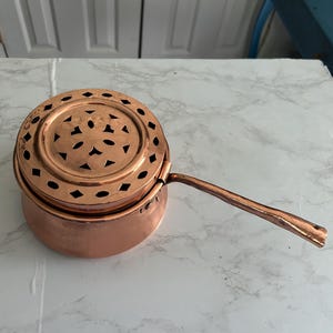 Op de afbeelding: Een koperen steelpan met een lange handgreep en een geperforeerd deksel. De pan is rond met een licht uitlopende basis. Het deksel heeft decoratieve uitgesneden vormen. De handgreep is aan de zijkant van de pan bevestigd.
