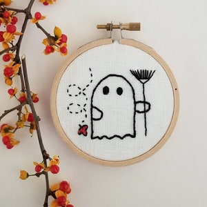 May include: Embroidered hoop art featuring a white ghost holding a flower with a red ladybug flying nearby. The hoop is made of wood and has a white fabric background.
