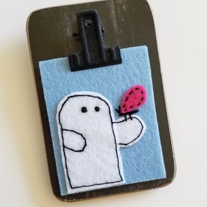 May include: A white felt ghost with black eyes holding a red felt ladybug on a blue felt background. The felt piece is attached to a wooden clipboard.