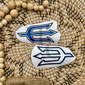 May include: Two white trident-shaped stickers with blue and black accents. The stickers are on a woven background with wooden beads. The trident design features a central arrow with two side arrows.