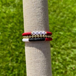 May include: Stacked bracelets with red, black, white, and gold beads. The top bracelet spells out the name "BOAZ" in white letter beads. The bracelets are displayed on a beige fabric roll against a green background.