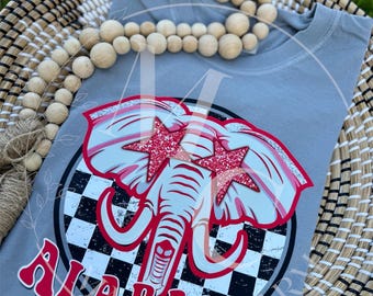 May include: A gray t-shirt featuring a graphic of an elephant with red star accents and the word "ALABAMA" in red. The design is set against a black and white checkered background. The shirt is displayed in a woven basket.