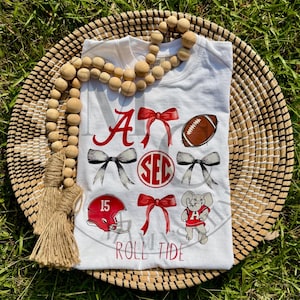 May include: White t-shirt with red and black graphics. Features a football, helmet, and elephant design. The text "A", "SEC", and "Roll Tide" are also visible. The shirt is displayed on a woven basket.