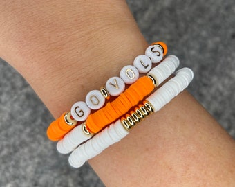 May include: A set of three bracelets featuring white beads with gold lettering spelling "GO VOLS" and orange beads.