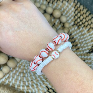 May include: Two bracelets featuring baseball-themed beads. One bracelet has white beads with red stitching, and the other has white disc-shaped beads. Gold-colored spacers separate the baseball beads. The bracelets are worn on a wrist.