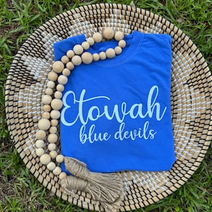 May include: A royal blue sweatshirt with the words "Etowah blue devils" in white script. The shirt is displayed on a woven basket with a wooden bead garland and a jute tassel.
