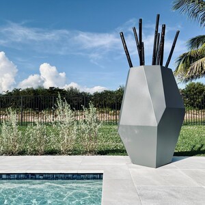 May include: A gray geometric vase with black sticks sticking out of the top. The vase is sitting on a gray patio next to a swimming pool.