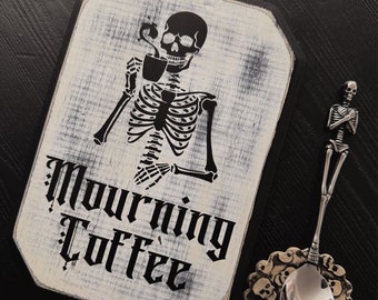 May include: A white wooden sign with black lettering that reads "Mourning Coffee". A black skeleton figure is depicted drinking from a coffee cup. The sign is on a black background.