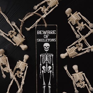 May include: A black wooden sign with white text that reads "Beware of Skeletons" and a white skeleton illustration. The sign is surrounded by multiple white plastic skeletons on a black background.