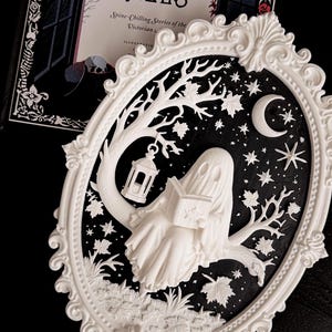 May include: An oval white frame containing a relief sculpture of a ghost sitting on a tree branch reading a book. The scene features a hanging lantern, maple leaves, stars, and a crescent moon against a black background. Behind it is a book titled Ghostly Tales.