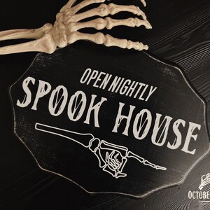 May include: Black wooden sign with white text that reads "Open Nightly Spook House". A white skeleton hand points to the right. The sign is on a black surface.