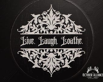 May include: A black round wooden sign with a white floral design and the text "Live. Laugh. Loathe."