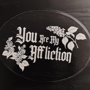 May include: A black circular wooden sign with white text and floral designs. The text reads "You are my affliction."