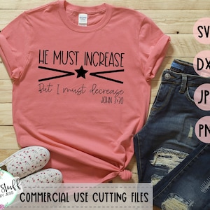 以下が含まれることがあります： 黒い文字で「HE MUST INCREASE But I must decrease JOHN 3:30」と書かれたピンクのTシャツ。テキストは白い背景にあり、黒い星と2本の黒い線で囲まれています。Tシャツは木製の表面に置かれ、ジーンズと白い靴のペアが置かれています。