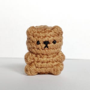 May include: A small, handmade crochet bear in a light brown colour. The bear has a simple design with black eyes and a small black stitched mouth. The bear is standing upright on a white surface against a white background.