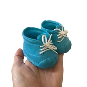 May include: A pair of small, turquoise-blue boots with white laces. The boots have a rounded toe and a textured sole. The laces are tied in a bow. The boots are held in a hand against a white background.