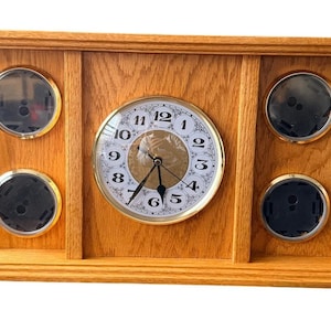 May include: Wooden clock with a light brown finish. The clock face has a white background with black numerals and gold-colored hands. It has a gold-colored bezel and two circular gauges on each side.