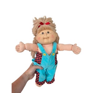 May include: A vintage Cabbage Patch Kid doll with blonde yarn hair and a red bow. The doll wears a turquoise corduroy overall with red and black checkered shorts. The doll has rosy cheeks and is in good condition.