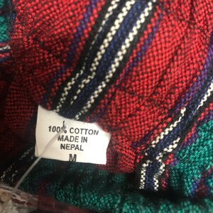 May include: A close-up of a red, green, and black plaid fabric with a white tag that reads "100% Cotton Made in Nepal M".