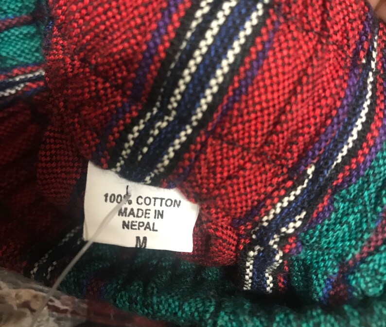May include: A close-up of a red, green, and black plaid fabric with a white tag that reads "100% Cotton Made in Nepal M".