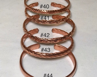 May include: Five copper bracelets, stacked on top of each other. The bracelets are all different sizes and have a braided design. The bracelets are labeled with numbers 40, 41, 42, 43, and 44. The text "Assorted" is written below the bracelets.