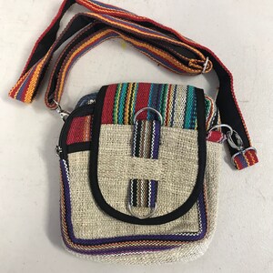 May include: A small, beige hemp crossbody bag with a colourful striped flap and a black strap. The bag has a zipper closure and two metal rings on the front.
