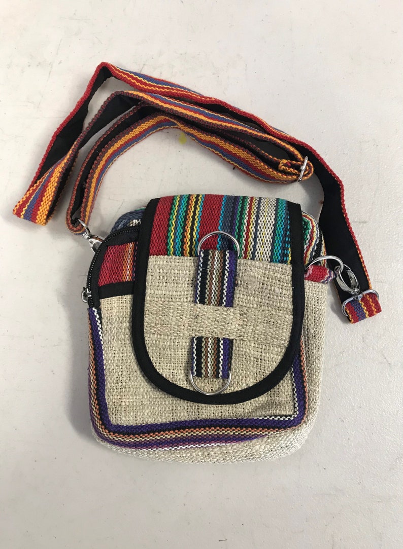 May include: A small, beige hemp crossbody bag with a colourful striped flap and a black strap. The bag has a zipper closure and two metal rings on the front.