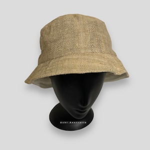 Puede incluir: Un sombrero de pescador beige hecho de tela tejida. El sombrero tiene una corona redondeada y un ala inclinada hacia abajo. Se muestra sobre una cabeza de maniquí negra sobre un fondo blanco. El texto "Achi Australia" es visible en la base.