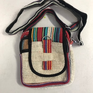 May include: A small, beige hemp crossbody bag with a black strap and colourful striped accents. The bag has a zipper closure and a flap with a metal ring.