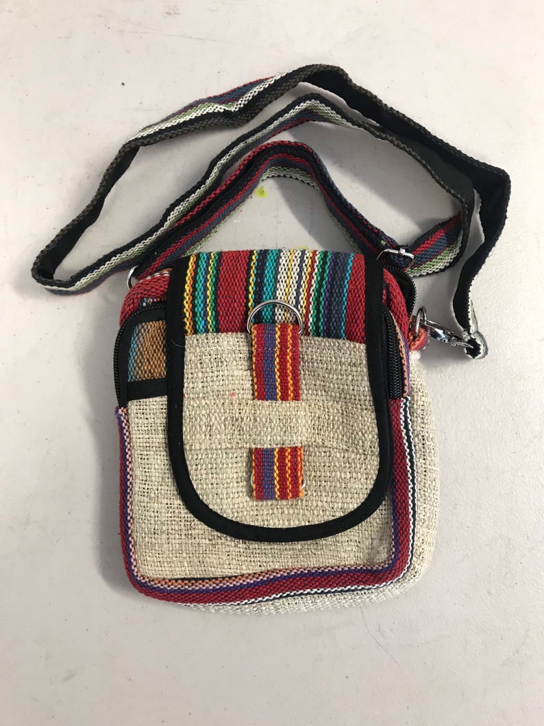 May include: A small, beige hemp crossbody bag with a black strap and colourful striped accents. The bag has a zipper closure and a flap with a metal ring.