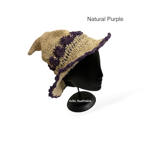 Puede incluir: Un sombrero de bruja morado natural hecho de paja tejida. El sombrero presenta una copa puntiaguda y un ala ancha con un borde festoneado. El ala y la copa están adornadas con un ribete morado oscuro y flores decorativas. El texto "Natural Purple" es visible.