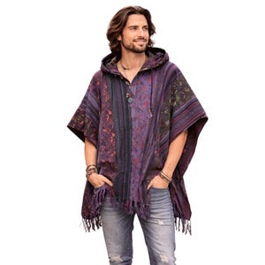 May include: A hooded poncho in shades of purple, blue, and green with a patterned design. The poncho has a fringed hem and a button closure at the neckline. The model is wearing blue jeans.