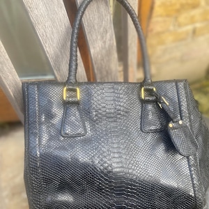 May include: Black leather handbag with a snakeskin effect. The bag features a structured rectangular design, a braided top handle, and gold-tone hardware. A small tag hangs from the side.