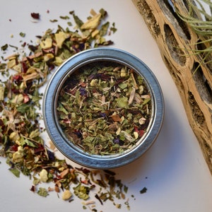 May include: A silver metal tin filled with a blend of dried herbs and flowers. The tin is surrounded by loose herbs and flowers on a white background.