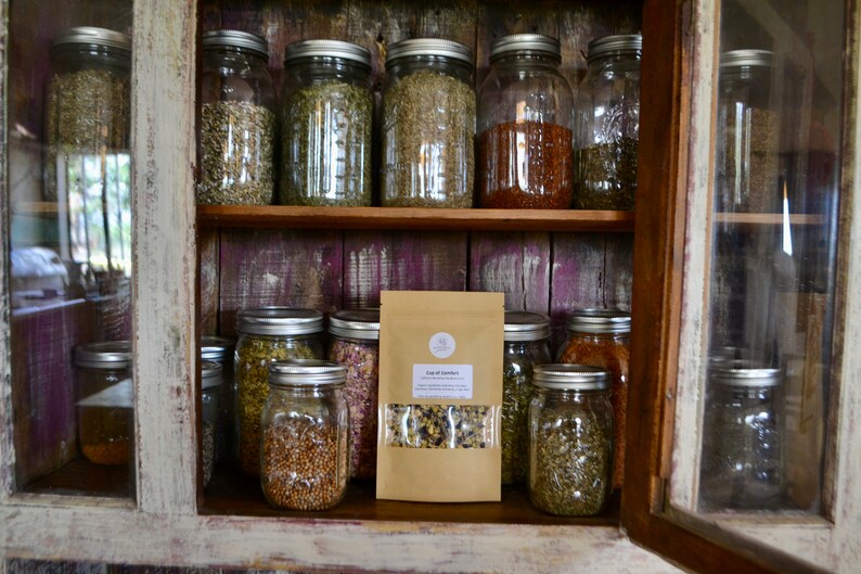 May include: A wooden cabinet with glass doors filled with glass jars containing dried herbs and spices. A brown paper bag with the text "Cup of Calm" is on the bottom shelf.