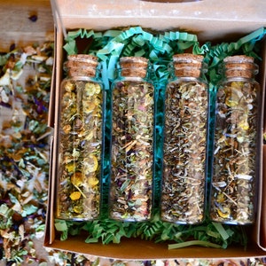 May include: Four small glass jars filled with dried herbs and flowers, arranged in a box with green shredded paper. The jars have cork lids.