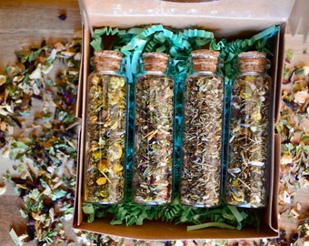 May include: Four small glass jars filled with dried herbs and flowers, arranged in a box with green shredded paper. The jars have cork lids.