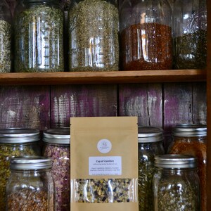 May include: A brown paper bag with a clear window showing a blend of dried herbs and flowers. The bag is labelled "Cup of Comfort" and lists the ingredients as "organic ingredients: Blackberry, Rose Buds, Rose Hips, Chamomile, Lavender, Lemon Balm, and a hint of warming spices for your tea."