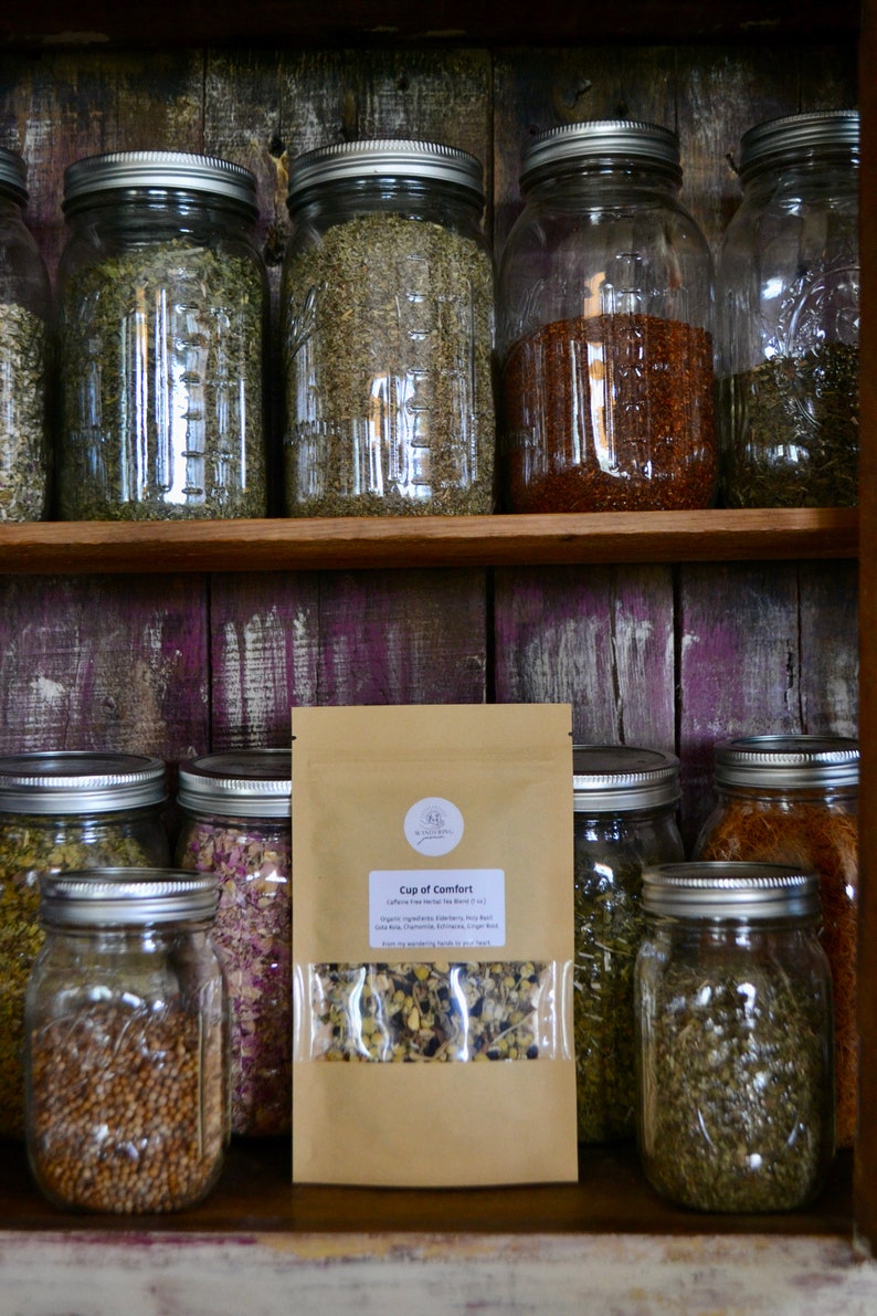 May include: A brown paper bag with a clear window showing a blend of dried herbs and flowers. The bag is labelled "Cup of Comfort" and lists the ingredients as "organic ingredients: Blackberry, Rose Buds, Rose Hips, Chamomile, Lavender, Lemon Balm, and a hint of warming spices for your tea."