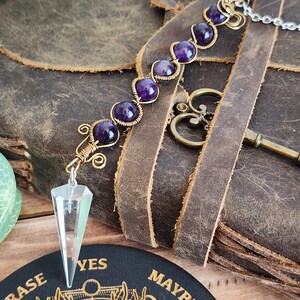 May include: A handcrafted pendulum with a clear crystal point and a chain. The pendulum features a series of purple amethyst beads wrapped in gold-colored wire. The background includes a leather journal and a decorative key.