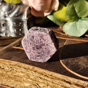 May include: A raw, hexagonal-shaped ruby crystal with a deep magenta and purple hue. The stone features a textured, crystalline surface with visible natural inclusions and a matte finish, showcasing its distinct geometric, six-sided structure.