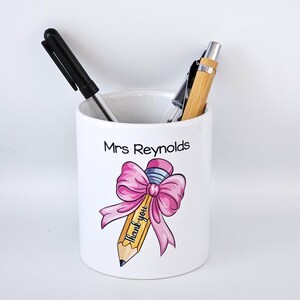 May include: White ceramic pencil pot with the name "Mrs Reynolds" and a pencil design featuring a pink bow and the words "Thank you". A black pen and a wooden pen are inside.