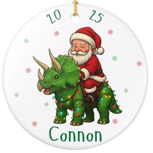 May include: A round white ceramic ornament featuring a green triceratops dinosaur wrapped in colorful Christmas lights with Santa Claus riding on its back. The text 20 25 is printed at the top and the name Connon is printed at the bottom.