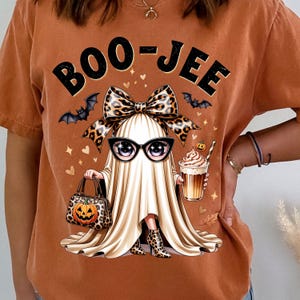 May include: A burnt orange t-shirt featuring a graphic of a ghost wearing black glasses and a leopard print bow. The ghost holds a leopard print handbag with a pumpkin design and a coffee drink. The text BOO-JEE is printed above the ghost.